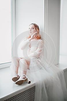 a gentle romantic young woman in a white transparent negligee with a long train is sitting on the windowsill in a bright