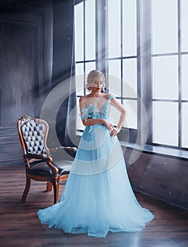 A gentle, snow queen stands by the window in a fabulous dress with bare shoulders. The room is filled with magical rays