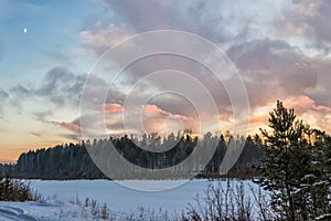 A gentle sunset over a forest and a snow-covered river. The new moon appeared in the sky at the same time as the sunset.