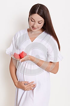 Gently pregnant woman with red booties, touching her belly, expectant mother with baby`s booties posing against white wall,