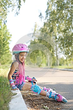 A genuinely laughing cheerful athletic girl on roller skates is sitting on the grass by the road.