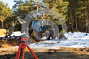 A geodesic level stands in front of a bulldozer for excavating a pit at a construction site. The excavator bucket digs a hole for