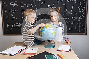 Geography lesson: A boy and a girl are sitting on a table and studying the globe on the background of a school board.