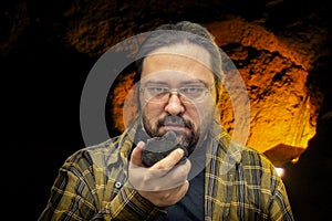 A geologist man in clean clothes with glasses holds in his hand a piece of coal on a blurred cave background.