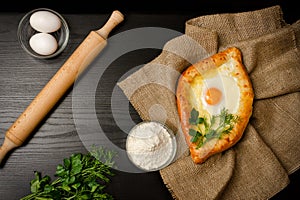 Georgian cuisine. Top view of khachapuri on sackcloth, flour, eggs and rolling pin. Black table