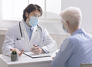 Geriatrician in medical mask talking with senior patient in office