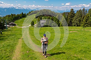 Gerlitzen - Woman with baby carrier on hiking trail along alpine meadow with scenic view of mountain range Julian Alps