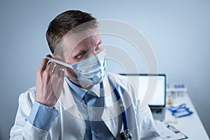 German European young doctor looking at camera wearing glasses and mask in clinic office at workplace with tired look