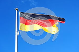 German flag with stripes in black, red, gold on a pole, is fluttering with frayed seam in the wind against a blue sky, copy space