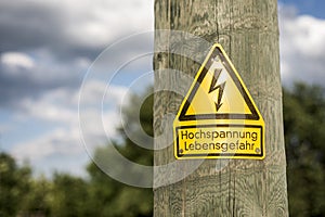 German high voltage sign mounted on wooden pole with green trees in background