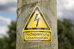 German high voltage sign mounted on wooden pole with green trees in background