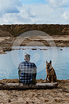 German shepherd with owner. Rear view. Young man with dreadlocks is sitting on wooden log on riverbank with dog enjoying views of