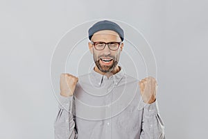 Gesture of success. Jubilant overjoyed unshaven man raises clenched fists, wears spectacles and formal shirt, celebrates his