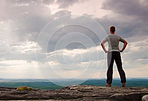 Gesture of triumph. Happy hiker in greyshirt and dark trousars. Tall man on the peak of sandstone cliff watching down to landscape