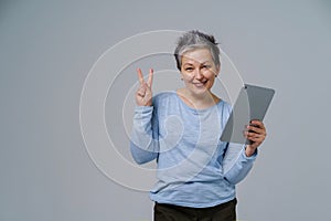 Gesturing V or victory mature grey haired woman in 50s holding digital tablet working or shopping online, checking on