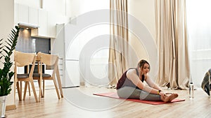 Get bent. Young curvy woman in sportswear using wireless earphones, exercising on a yoga mat at home