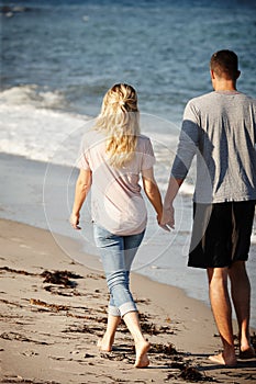 Getting away from it all. Rearview shot of a young couple walking hand in hand along a sandy beach.