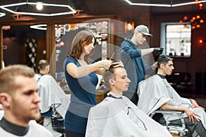 Getting haircut. Side view of professional barber girl holding scissors and comb in hands and cutting hair of young guy