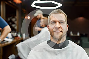 Getting new haircut. Portrait of young handsome bearded man sitting in barbershop chair and smiling at camera. Visiting