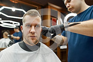 Getting new trendy haircut. Professional barber working with hair clipper, serving young handsome man sitting in