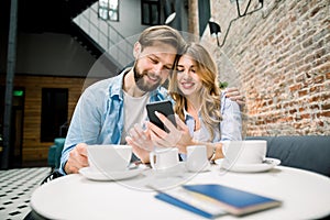 Getting ready for the trip with a coffee. Happy couple sitting at the table, drinking coffe or tea, using phone