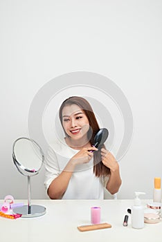 Getting rid of tangles. Beautiful young woman looking at her reflection in mirrorand brushing her long hair while sitting at the