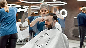 Getting trendy haircut. Young professional barber girl, female hairdresser working with hair clipper, serving handsome