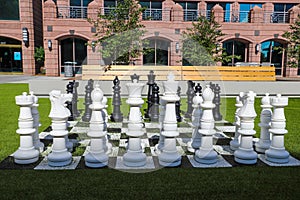 A giant black and white chess set on lush green grass surrounded by lush green and autumn colored trees near buildings