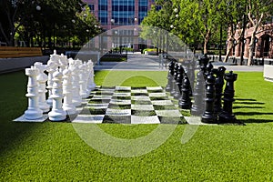 A giant black and white chess set on lush green grass surrounded by lush green and autumn colored trees near buildings