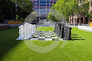 A giant black and white chess set on lush green grass surrounded by lush green and autumn colored trees near buildings