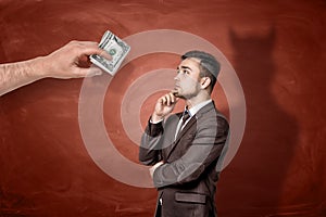 A giant hand with a roll of dollar bills in front of a thinking businessman who cast a shadow of a devil. photo