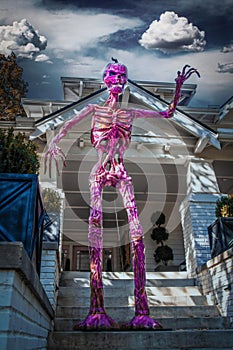 Giant purple skeleton monster with pumpkin head stands on steps  towering over vintage home decorated for Halloween