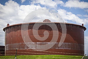 Giant rusty oil tank in tank farm in Cushing Oklahoma behind barbed wire fence with pretty blue sky background