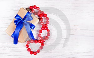 Gift box with blue bow and red coral beads on a white wooden background. Surprise to the woman.