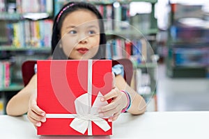 Gift box for kids girl. White box with red bow in the smiling asian girl hands for give a gift in the library, happy and surprise