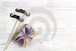 Gift box with rainbow LGBT ribbon and pair of decorative mustache on a light wooden background