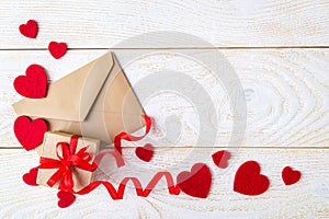 Gift box with red ribbon bow, brown paper envelope and valentine hearts on a white wood textured surface. Greeting card with copy