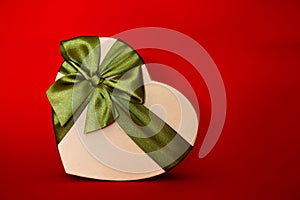 Gift box in the shape of a heart tied with a green bow on a red background. New Year`s packaging