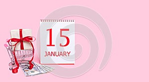 A gift box in a shopping trolley, dollars and a calendar with the date of 15 january on a pink background.
