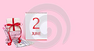 A gift box in a shopping trolley, dollars and a calendar with the date of 2 june on a pink background.