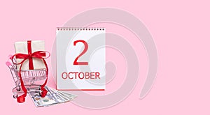 A gift box in a shopping trolley, dollars and a calendar with the date of 2 october on a pink background.