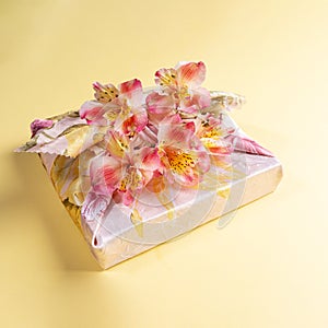 Gift box trendy wrapped in floral fabric in japanese Furoshiki technique with flowers Alstroemeria on yellow backdrop.