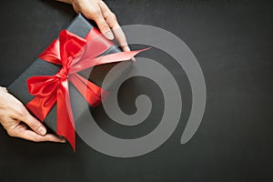 Gift box wrapped in black paper with red ribbon in female hand on black surface.