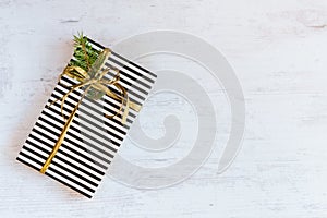 Gift box wrapped in black and white striped paper with golden ribbon and fir branch on a white wooden background. Christmas concep