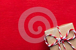 Gift box wrapped in brown Kraft paper tied with striped ribbon on crimson red textured background. Trendy Christmas New Years card