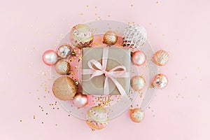 Gift box wrapped in craft paper with pink ribbon on pale pink background with glitters and golden Christmas tree balls