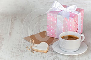 Gift box wrapped in pink dotted paper, heart shaped love cookie, a cup of coffee and an empty kraft card over a white wood