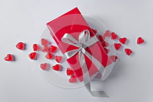 Gift-box is wrapped in red paper with silver ribbon bow. Isolated on a white background in a pile of jelly hearts.