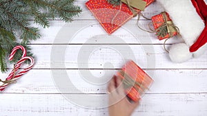 A gift box wrapping in the red paper appears on wooden background, and female hand picks up a present. Flat lay. Christmas decorat