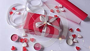 Gift box wrapping in a red paper with white ribbon bow on the white background with romantic decor: candles, red hearts.